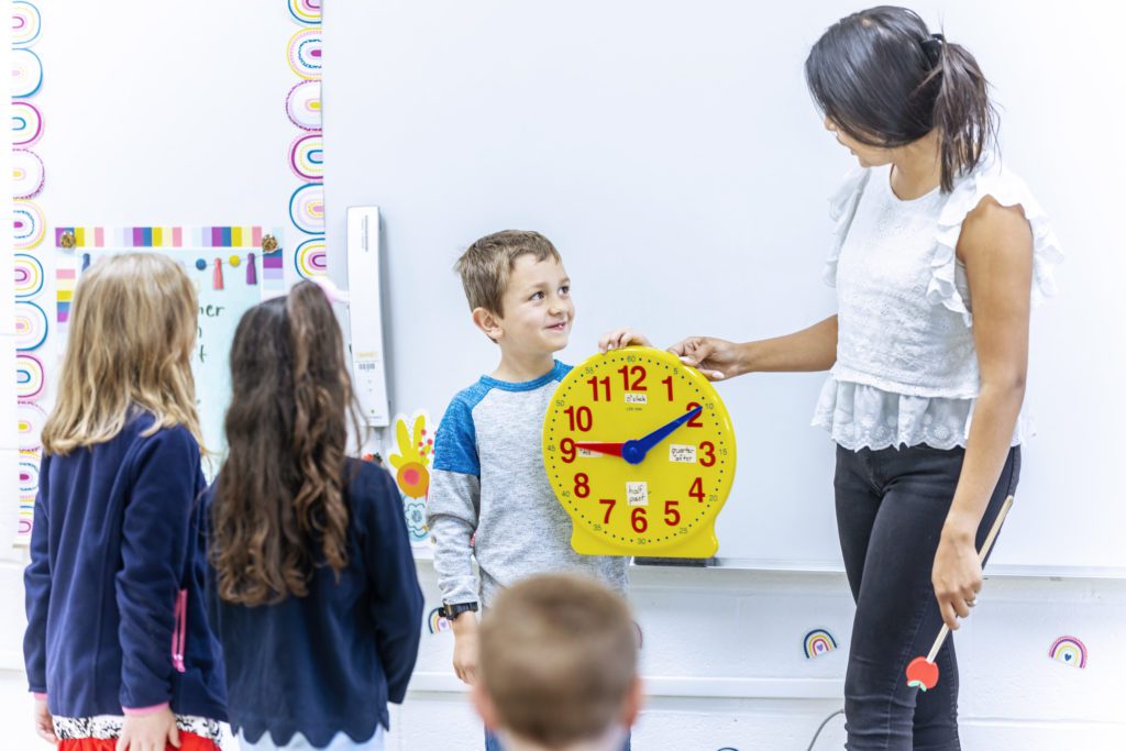 Young boy holding a clock, smiling at his teacher in a classroom